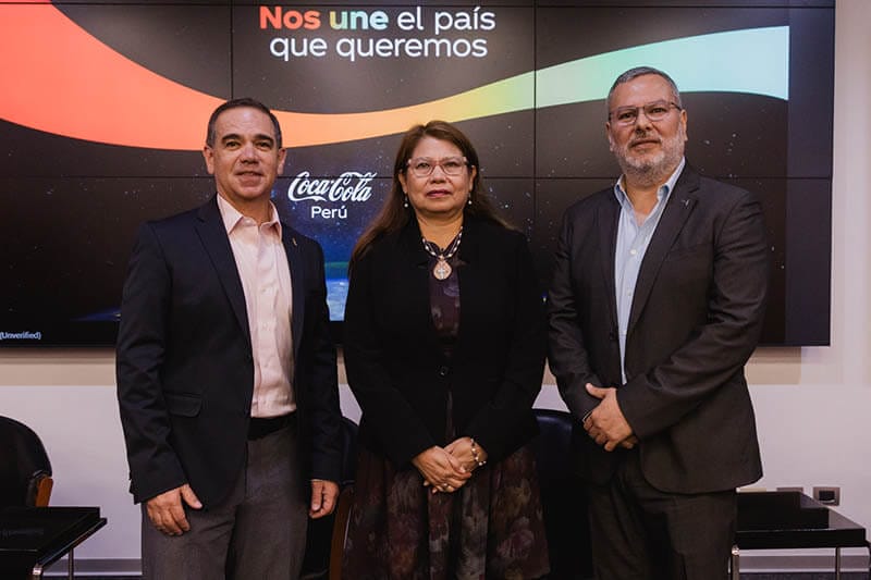 Tres personas posando frente a una pantalla con el logo de Coca-Cola y el mensaje 'Nos une el país que queremos'.