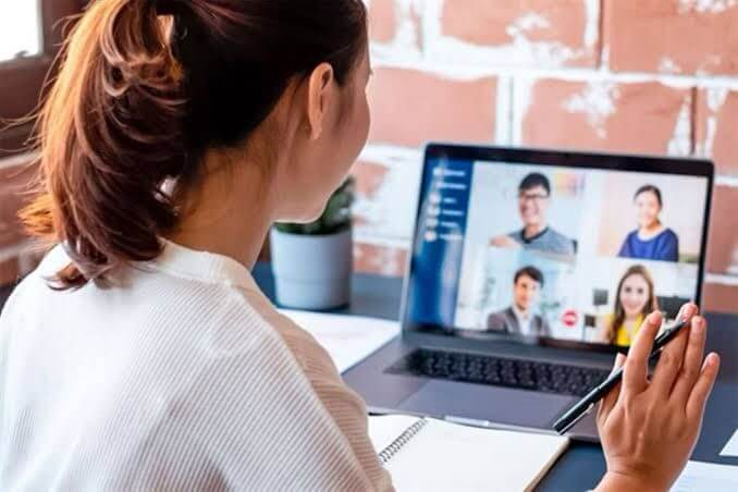 Mujer participando en una videoconferencia desde una laptop, con varias personas visibles en la pantalla y tomando notas con un bolígrafo.