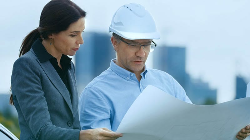 Una mujer y un hombre revisan planos de construcción al aire libre, con edificios de fondo. El hombre lleva un casco de seguridad y gafas, mientras que la mujer viste un traje formal.