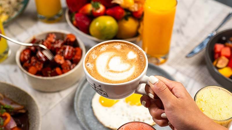 Una mano sostiene una taza de café con un diseño de corazón en la espuma, rodeada de varios platos de comida, incluyendo frutas, batidos y huevos, sobre una mesa bien decorada.