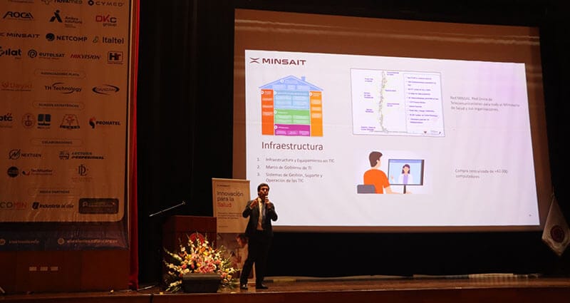 Presentación de Erick Cortez de Minsait en un evento sobre innovación en salud, con un gráfico de infraestructura y sistemas de TI en el fondo.