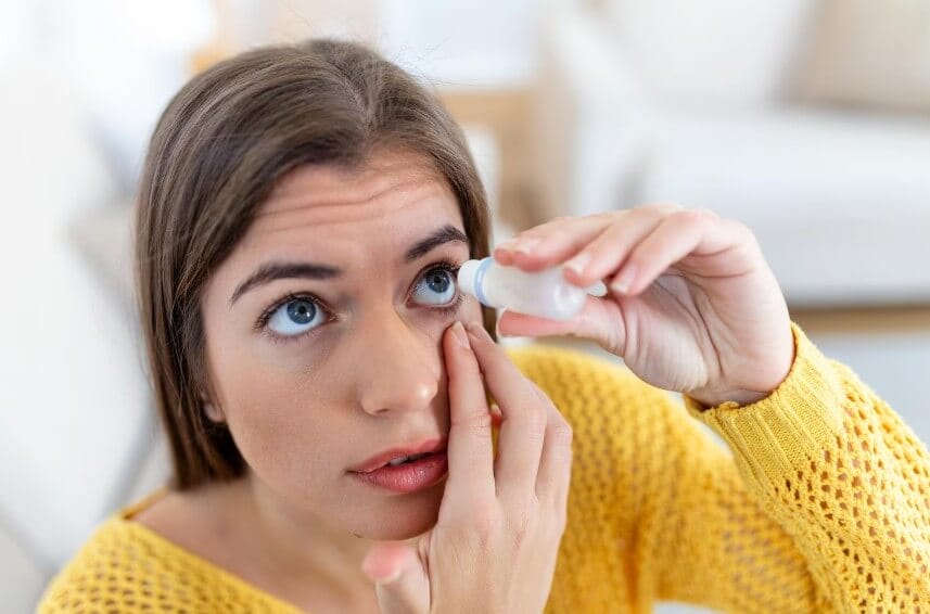 Mujer aplicando gotas para los ojos mientras sostiene el párpado en un ambiente interior, vestida con un suéter amarillo.