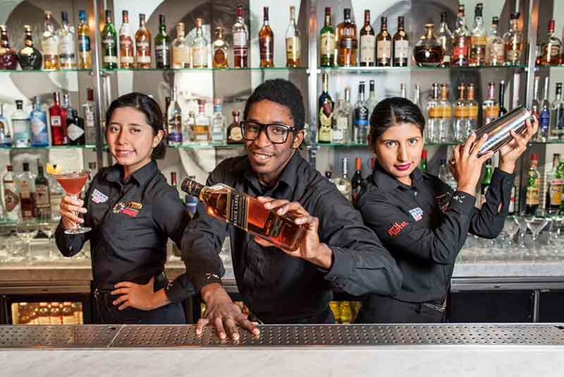 Tres bartenders posando detrás de una barra, uno sosteniendo una botella de licor y dos preparando cócteles, con estantes llenos de botellas de bebidas al fondo.