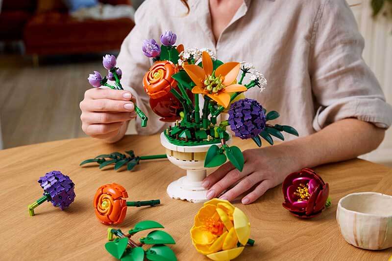Una persona armando un ramo de flores de LEGO en una mesa, rodeada de diversas piezas decorativas.