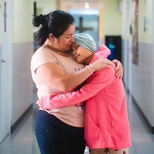 Una mujer abraza a una niña con un sombrero de color verde en un pasillo, mostrando un momento de ternura y apoyo emocional.
