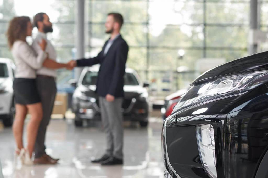Pareja negociando un contrato de leasing vehicular en un concesionario de automóviles, con un vehículo negro en primer plano.