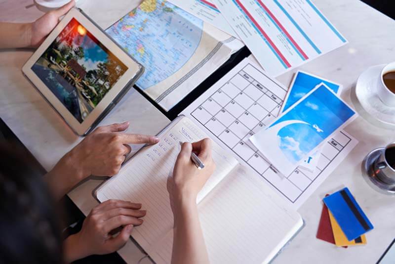 Una persona señalando información en una tablet mientras otra persona escribe en un cuaderno, rodeados de documentos de planificación de viajes, un mapa y tarjetas de pago sobre una mesa.