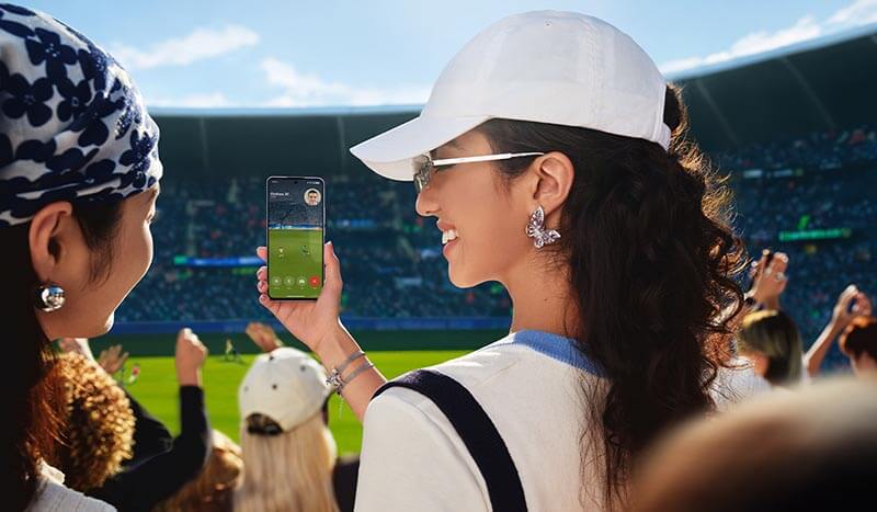 Mujer usando un smartphone para ver un evento deportivo en un estadio, con un ambiente festivo y seguidores en el fondo.
