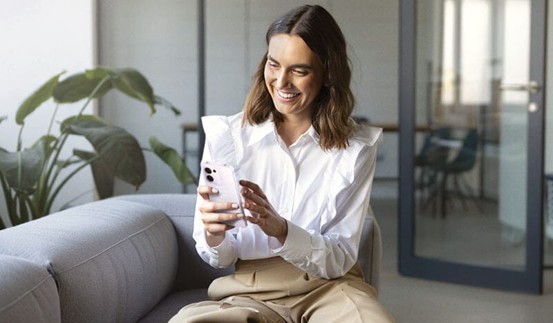 Mujer sonriente sentada en un sofá, usando un smartphone en un entorno moderno y luminoso.