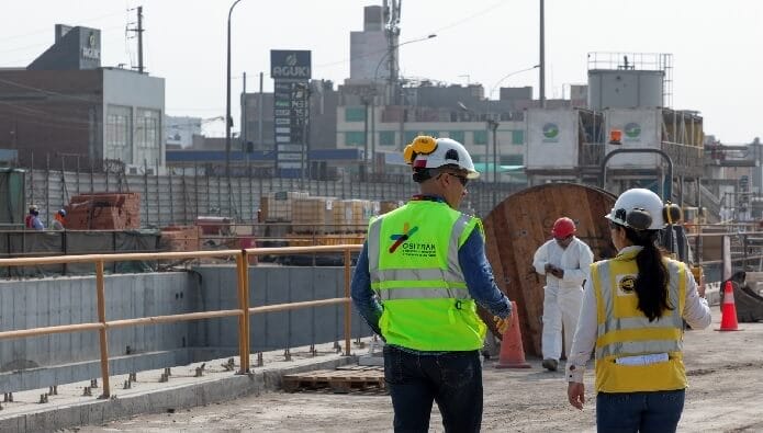Dos trabajadores con chalecos de seguridad y cascos en un sitio de construcción en Lima, supervisando el progreso de la obra, con maquinaria y otros trabajadores al fondo.