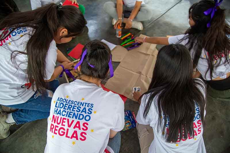 Grupo de adolescentes trabajando en taller de empoderamiento sobre salud menstrual, vestidos con camisetas que dicen 'Adolescentes Lideresas Haciendo Nuevas Reglas'.
