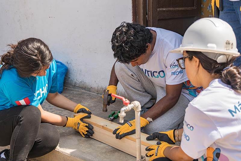 Tres personas trabajan juntas en un proyecto de instalación de soluciones de agua en una comunidad, utilizando herramientas y materiales como un martillo y una tabla de madera.