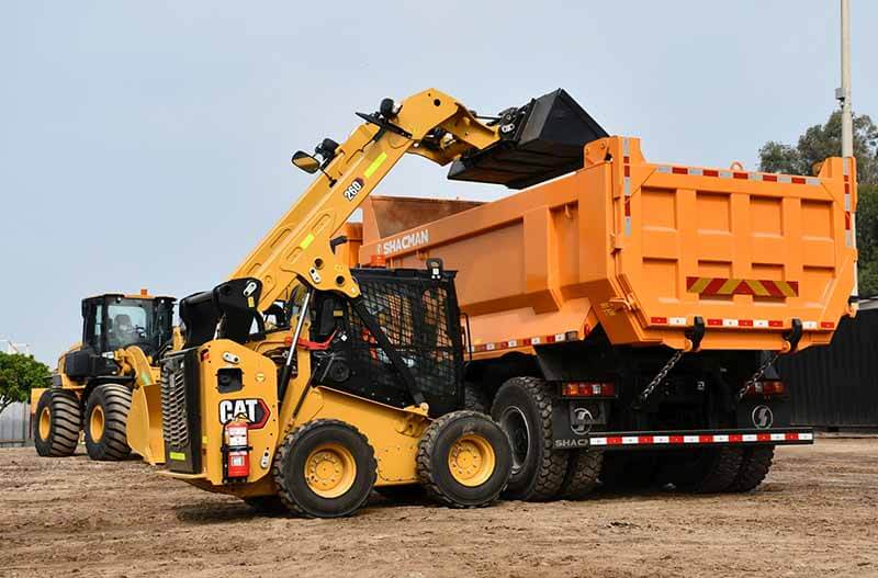 Minicargador Cat 260 levantando material en un sitio de construcción, con un camión de volquete naranja estacionado, mientras un minicargador Cat 250 se observa en el fondo.