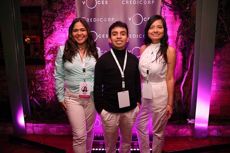 Tres jóvenes sonrientes posando juntos frente a un fondo decorado con luces moradas y el logotipo de VOCES y Credicorp.