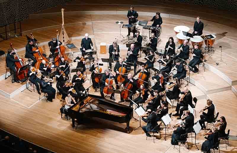 Orquesta Mahler en pleno ensayo con la pianista Yuja Wang interpretando en un escenario de madera, con una amplia variedad de instrumentos.