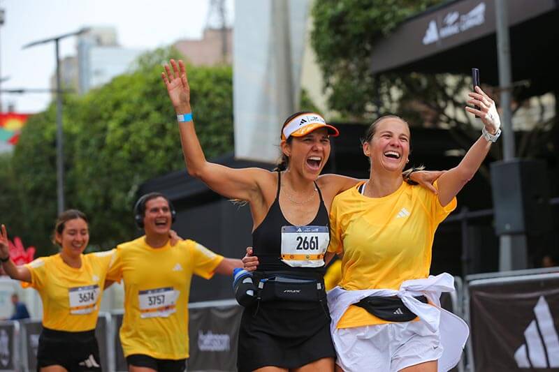 Dos mujeres corriendo y sonriendo durante una maratón, rodeadas de otros corredores y un entorno urbano.