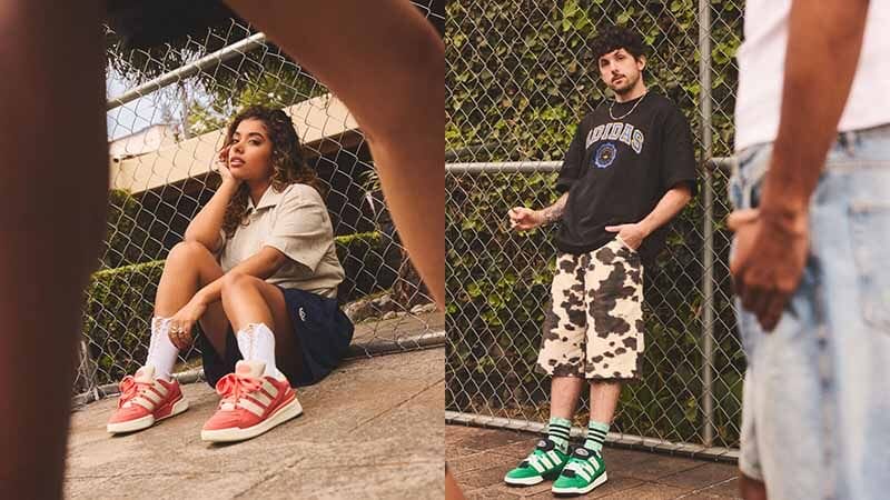 Una mujer sentada en el suelo, luciendo zapatillas rojas con calcetas, una camiseta gris y un short azul, posando de forma relajada. A la derecha, un hombre de pie, vistiendo una camiseta negra y shorts de camuflaje, con zapatillas verdes y calcetines de rayas, junto a una malla metálica.