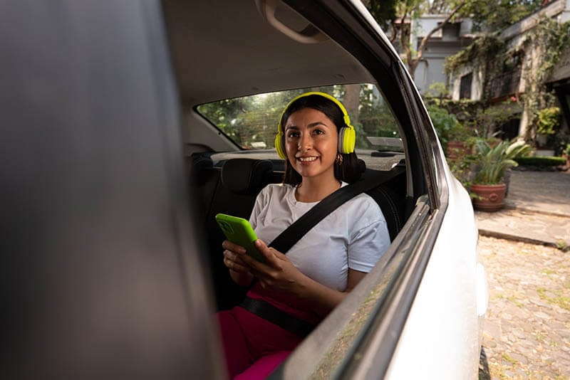 Mujer sonriendo en un auto, usando auriculares amarillos y sosteniendo un teléfono verde, mientras está sentada con el cinturón de seguridad puesto.