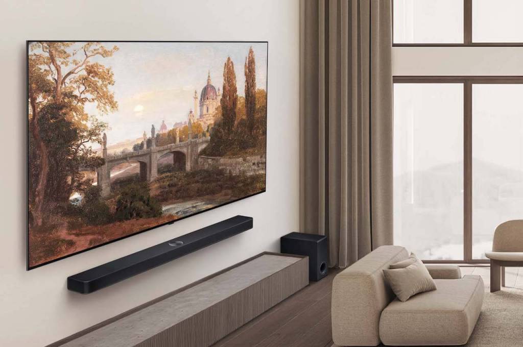 Televisor OLED montado en la pared mostrando una pintura paisajística, junto a una barra de sonido y un sofá en un salón moderno y luminoso.
