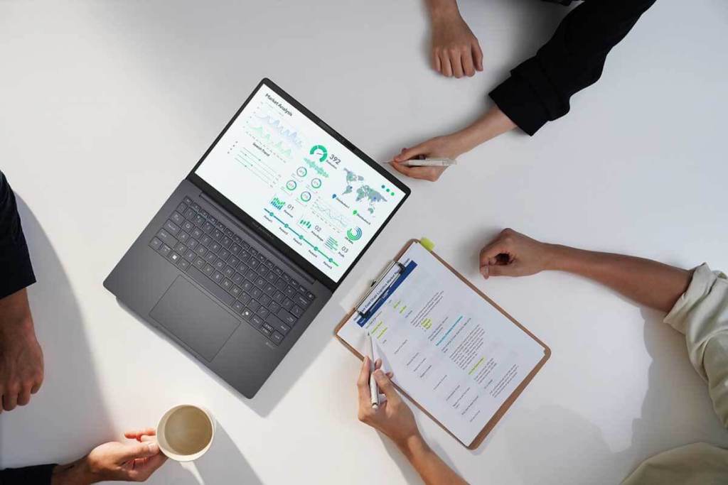 Una laptop ASUS ExpertBook B5 abierta sobre una mesa, mostrando gráficos de análisis de mercado en la pantalla, rodeada de manos de personas que sostienen un cuaderno y una taza de café.