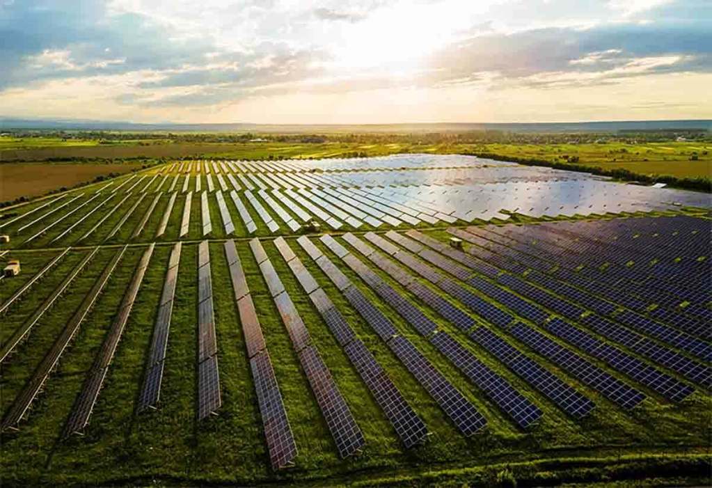 Panorámica de un extenso campo con paneles solares dispuestos en filas, capturando la luz del sol durante el atardecer, rodeado de paisajes agrícolas.