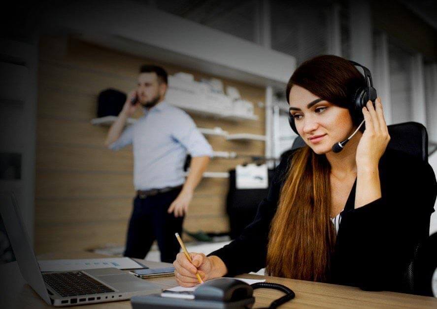 Mujer en un centro de atención al cliente usando un auricular y tomando notas, mientras un hombre hablando por teléfono está al fondo.
