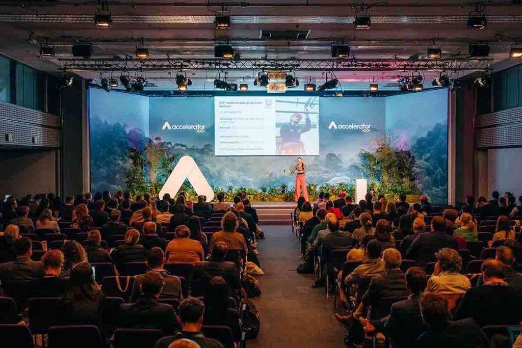 Evento de la Aceleradora 100+ con una presentadora en el escenario, dirigida a una audiencia en un entorno decorado que enfatiza la sostenibilidad.