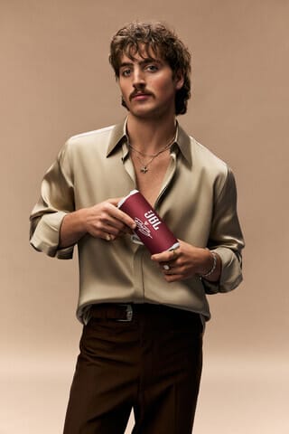 Joven artista masculino sosteniendo un altavoz JBL frente a un fondo neutro, vistiendo una camisa clara y pantalones oscuros.