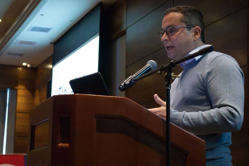 Un ponente hablando en un evento sobre ElevaTech, con un micrófono a su lado y una presentación en la pantalla detrás.