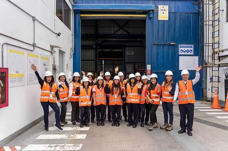 Grupo de estudiantes de secundaria en el Taller de Reparación de Transformadores de Hitachi Energy, vistiendo chalecos naranjas y cascos blancos, posando con entusiasmo.