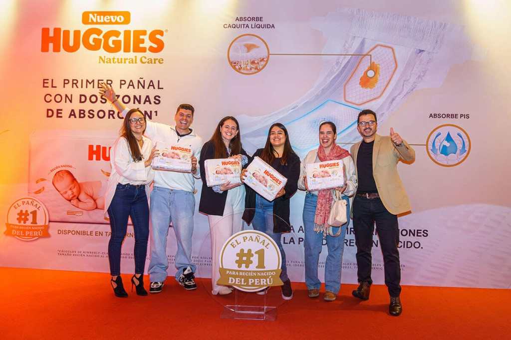Grupo de personas sonriendo y sosteniendo paquetes de pañales Huggies Natural Care durante un evento de lanzamiento, con un fondo que destaca el producto y su tecnología.