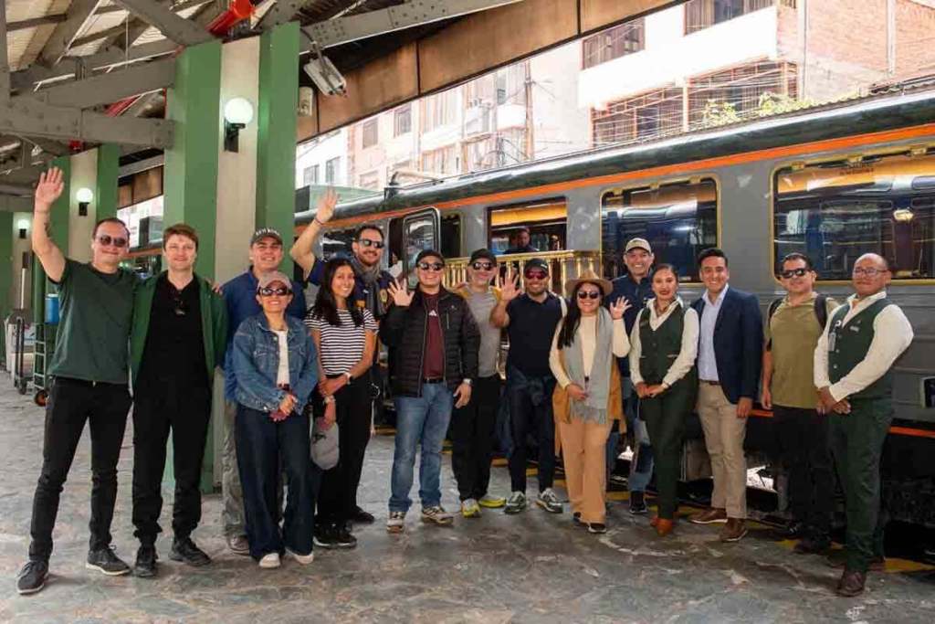 Un grupo de inversores y representantes turísticos sonríen y saludan frente a un tren de Inca Rail en una estación, destacando la experiencia de viajar hacia Machupicchu.