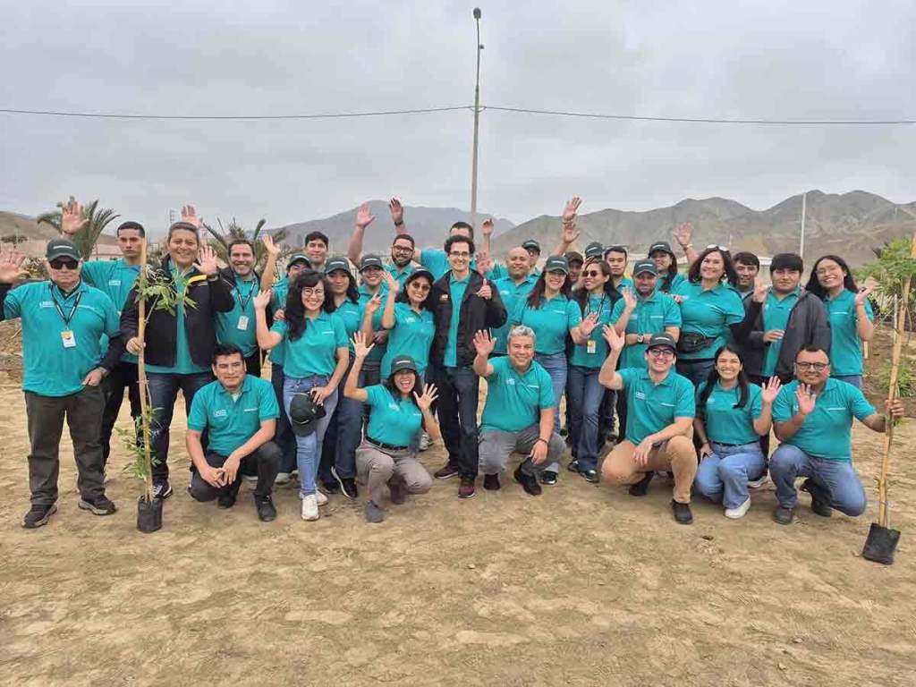 Grupo de personas sonrientes con camisetas verdes levantando las manos durante una jornada de arborización en un área al aire libre.