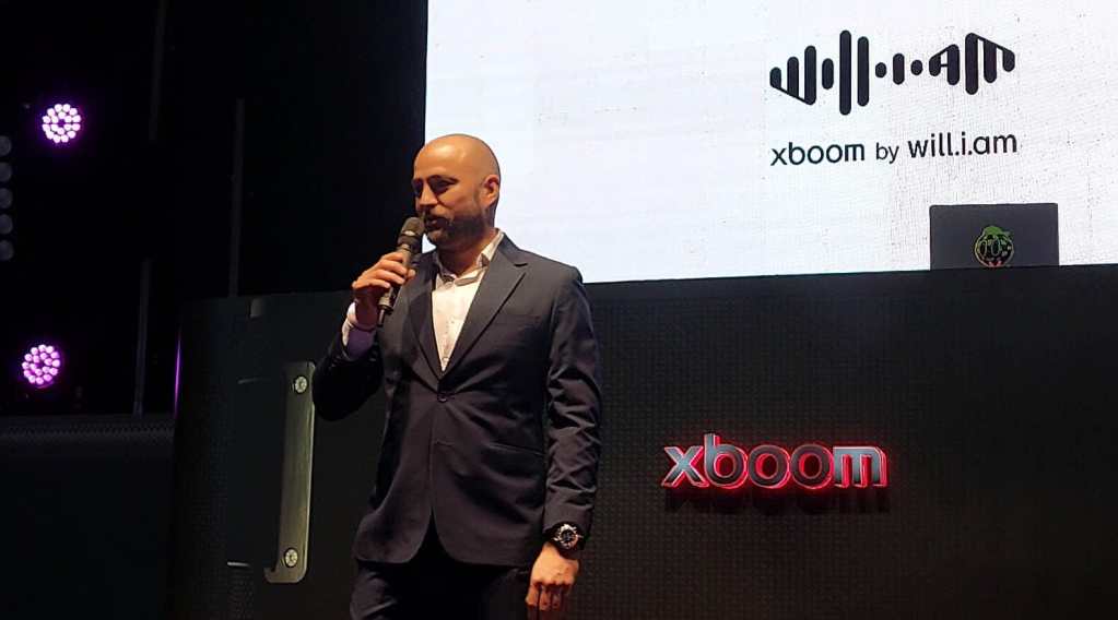 Presentador en evento de lanzamiento de la línea de audio xboom by will.i.am, con pantalla de fondo y logo del producto.