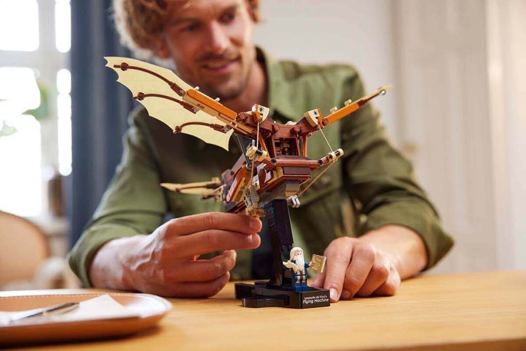 Un adulto ensamblando un set de construcción de un modelo de máquina voladora, con un minifigura junto a él, sobre una mesa en un ambiente acogedor.