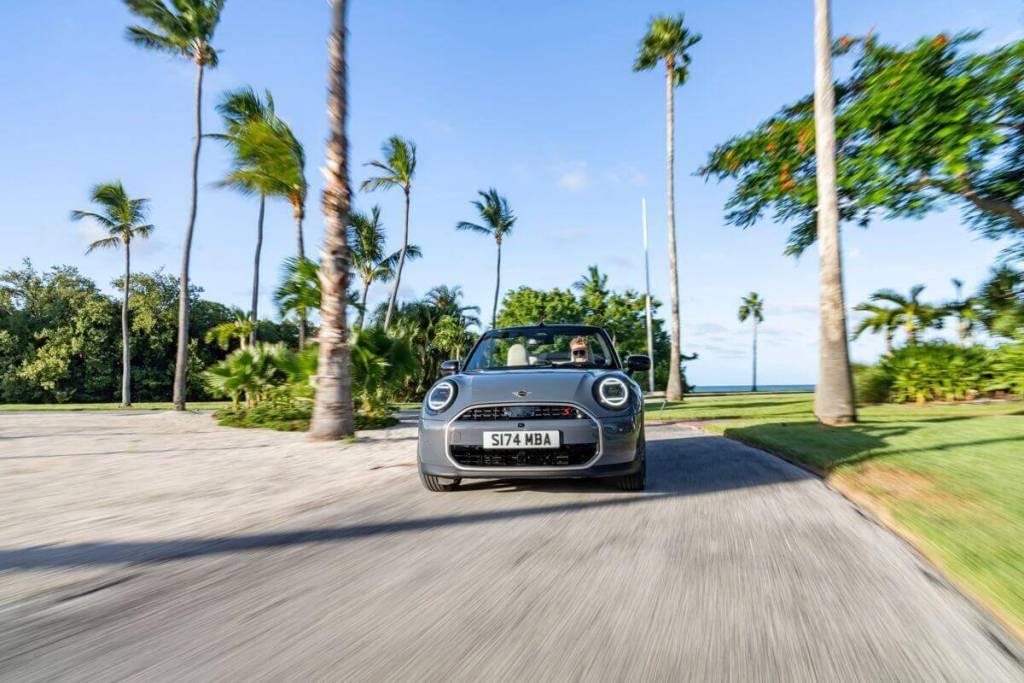Un MINI Cooper Convertible color gris circulando por una carretera rodeada de palmeras y vegetación exuberante, con el techo abierto y una persona al volante disfrutando del paisaje.