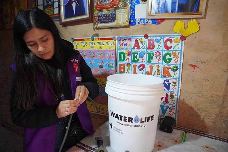 Una mujer está preparando un filtro de agua de la organización WATERisLIFE en un entorno comunitario, con un fondo que muestra un cartel didáctico con letras y colores.