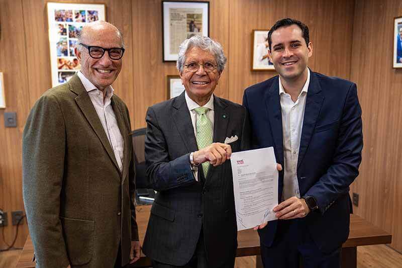 Tres hombres sonrientes posando juntos en una oficina, sosteniendo un documento, con fotografías enmarcadas en la pared de fondo.