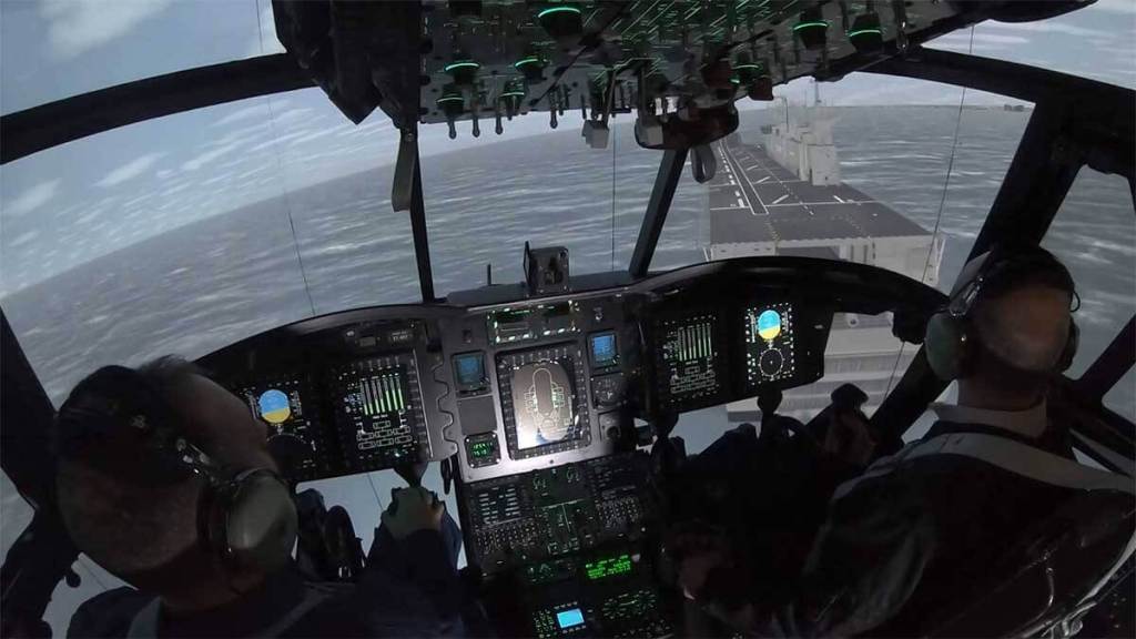 Vista de un simulador de vuelo dentro de una cabina, mostrando dos pilotos operando controles mientras se simula un paisaje marítimo y una embarcación en el horizonte.