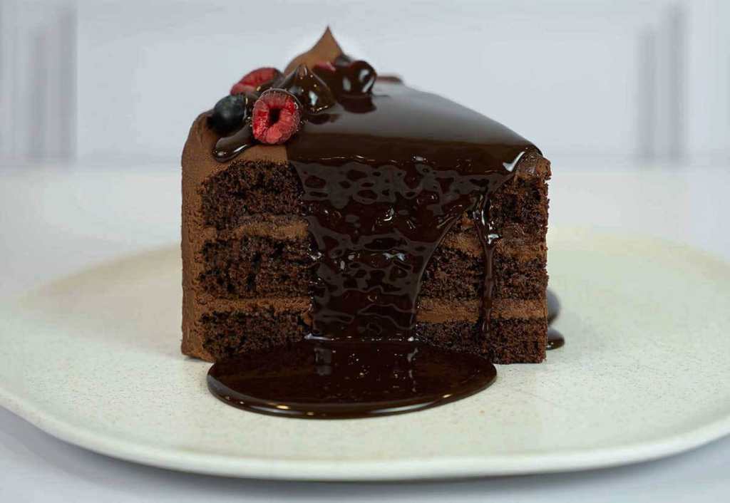 Una porción de torta de chocolate con capas visibles, cubierta de ganache brillante y decorada con frutas frescas. La salsa de chocolate se derrama sobre el plato.