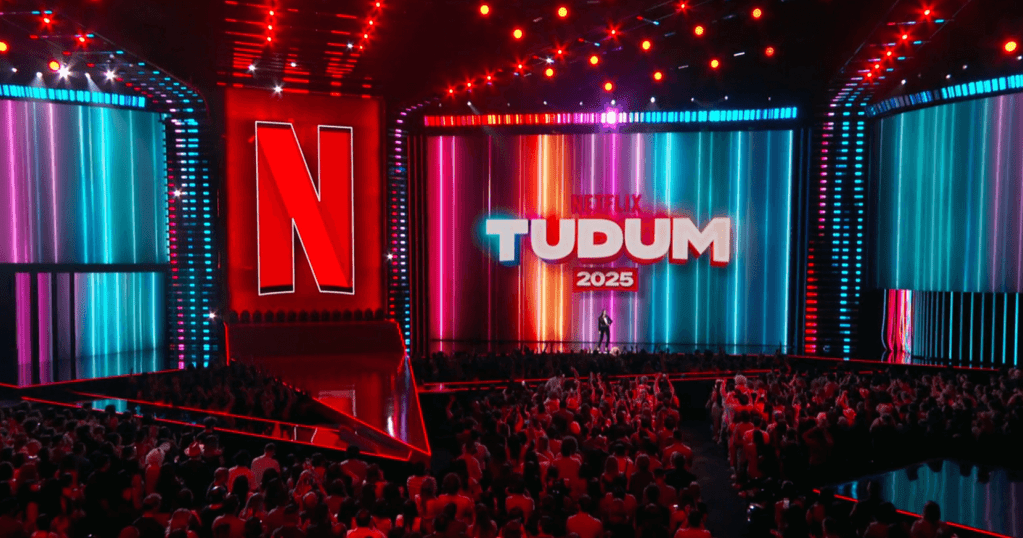 Escenario iluminado del evento TUDUM 2025 de Netflix, con un gran fondo que muestra el logotipo de Netflix y el texto 'TUDUM 2025', rodeado de una multitud animada.