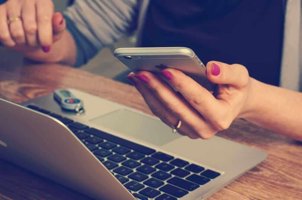 Una persona sostiene un teléfono móvil mientras está sentada frente a un ordenador portátil en una mesa de madera.