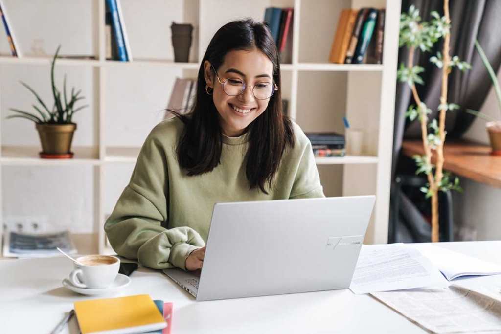 Mujer sonriente usando una laptop en un escritorio con una taza de café y material de estudio, rodeada de estanterías con libros y plantas.