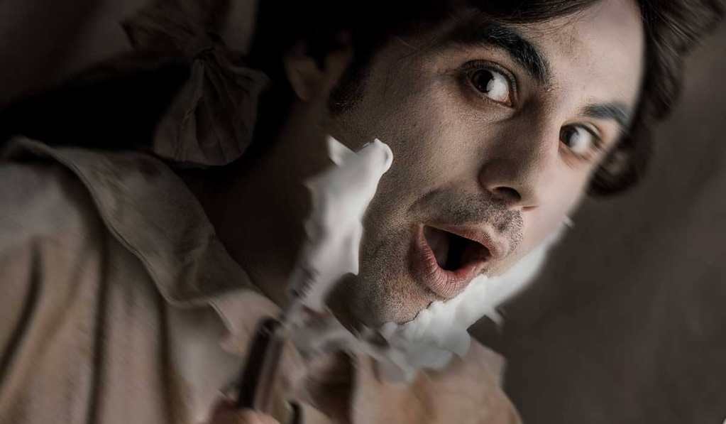 Primer plano de un hombre sorprendido con espuma de afeitar en la cara, sosteniendo una navaja de barbero, en un ambiente que evoca la Sevilla del siglo XVIII.