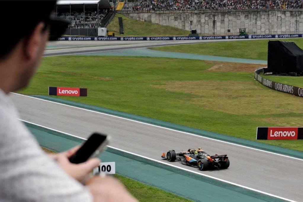 Un espectador observa una carrera de Fórmula 1 desde una tribuna, mientras un automóvil de carrera pasa por la pista. Se pueden ver patrocinadores como Lenovo en la pista y una multitud de espectadores en el fondo.