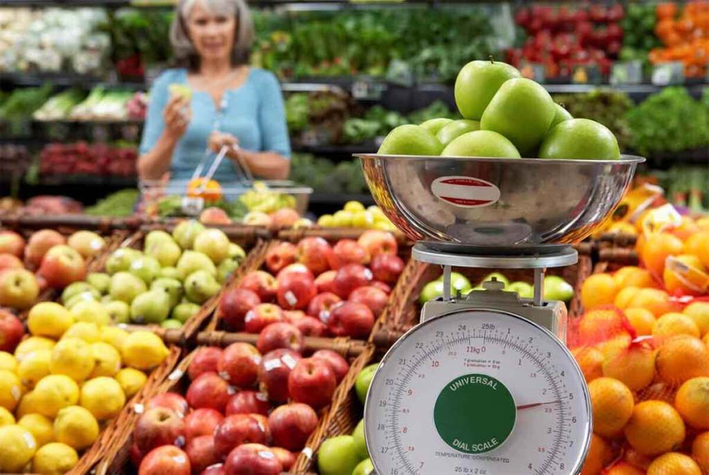 Una balanza con manzanas verdes en primer plano, en un mercado lleno de frutas coloridas, mientras una mujer escoge frutas al fondo.