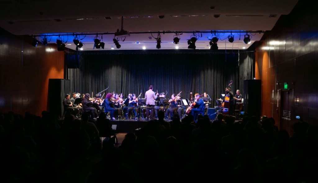 Orquesta Sinfónica del BRITÁNICO en un escenario, con músicos tocando diferentes instrumentos frente a una audiencia.