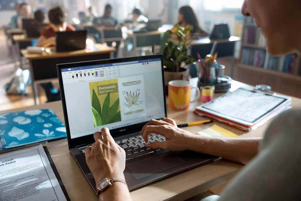 Un docente utilizando una laptop para crear contenido en un aula escolar, con estudiantes trabajando en sus propias computadoras al fondo.