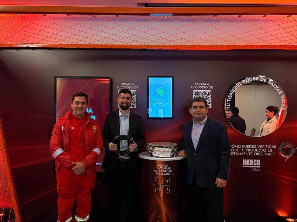 Tres hombres posan en un evento sobre la seguridad eléctrica, uno de ellos en uniforme de bombero. Al fondo, pantallas y un letrero que invita a escanear un código QR para verificar productos eléctricos.