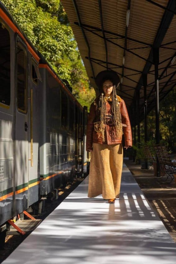 Modelo caminando por una pasarela en un tren, con un vestuario de alta moda, rodeada de un ambiente natural y ferroviario.